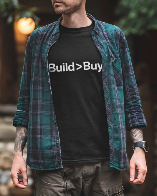 Person wearing a black t-shirt with 'Build > Buy' text and a plaid shirt outdoors.
