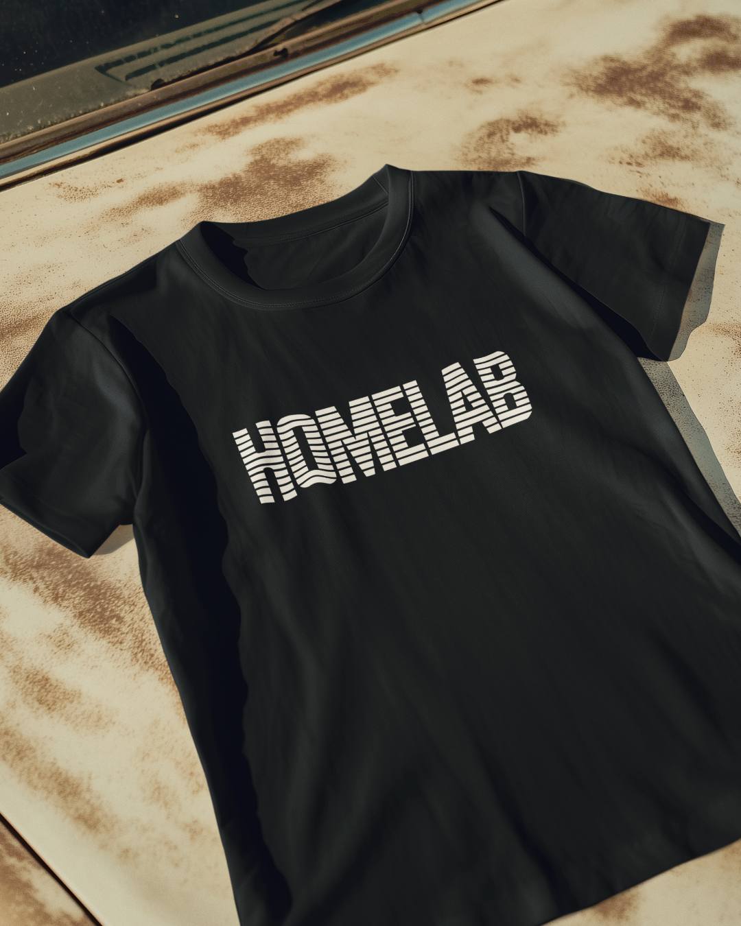 Black t-shirt laid in a row featuring a bold white “HOMELAB” wordmark with horizontal stripe/scanline effect; retro computer aesthetic with soft natural shadows.