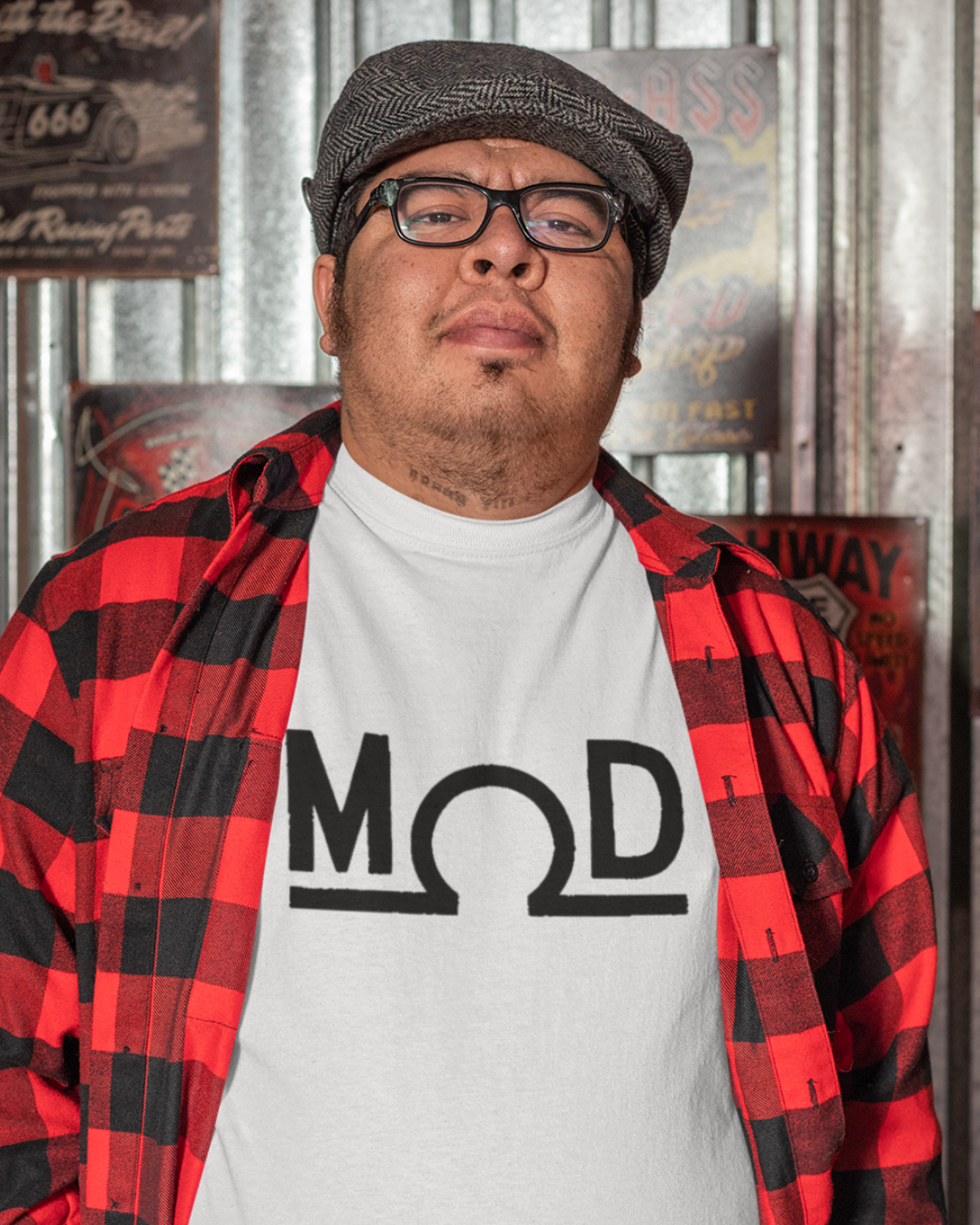Person in a red-and-black flannel over a white t-shirt featuring a bold black “MOD” wordmark with the Ω (ohm) symbol as the “O” and a solid baseline; photographed in a retro garage with metal wall panels and vintage signs.