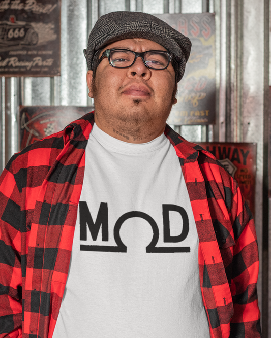 Person in a red-and-black flannel over a white t-shirt featuring a bold black “MOD” wordmark with the Ω (ohm) symbol as the “O” and a solid baseline; photographed in a retro garage with metal wall panels and vintage signs.