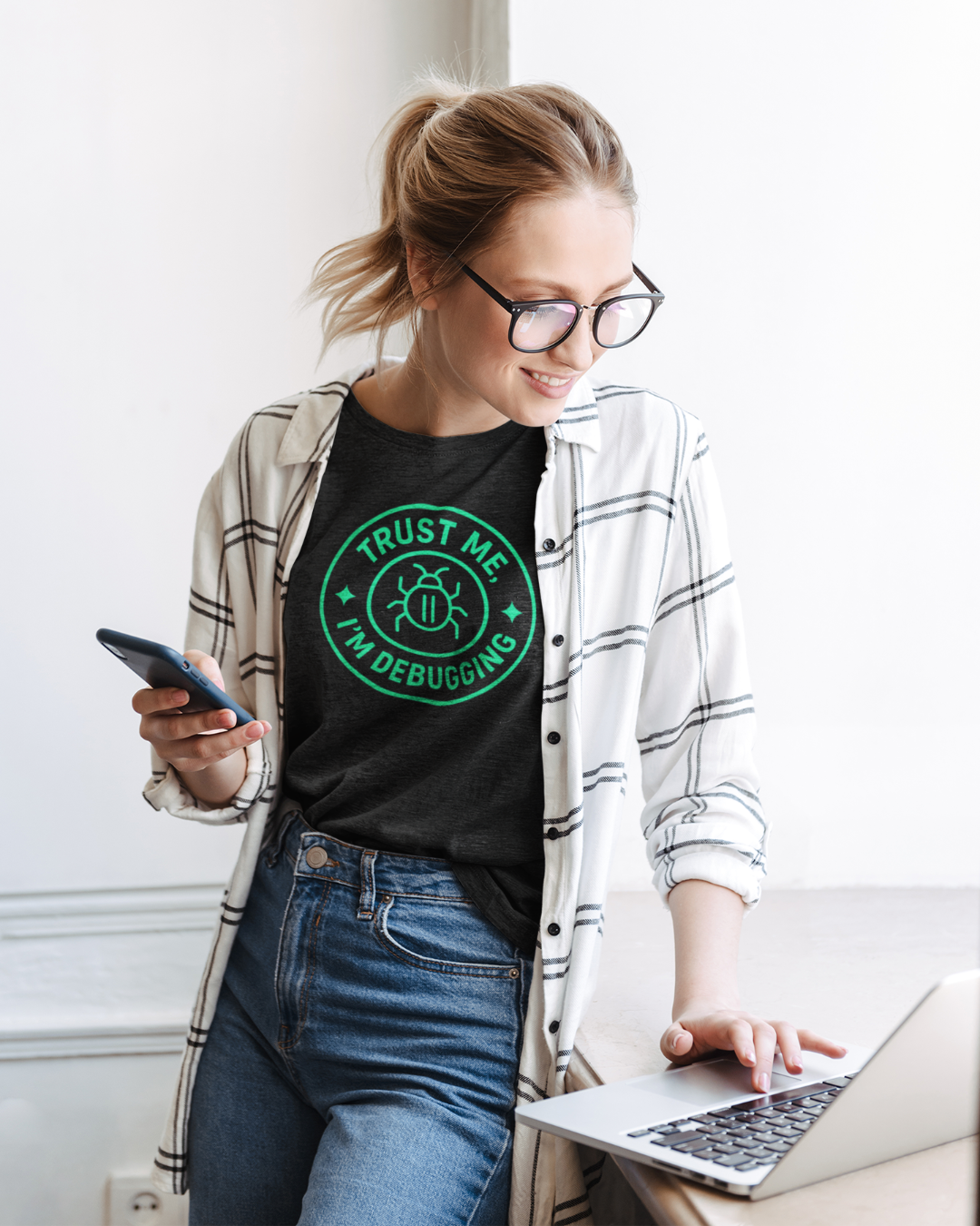 Woman wearing a black t-shirt with 'Trust Me, I'm Debugging' design, using a laptop.